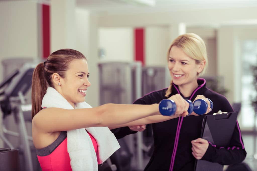 Hymyilevä personal trainer ohjaa naisasiakasta käsipainoharjoituksessa kuntosalilla
