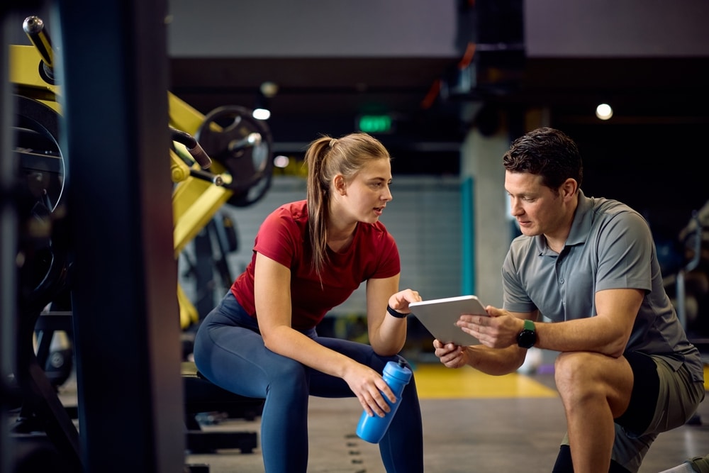 Personal trainer näyttää asiakkaalle harjoitusohjelmaa tabletilta modernissa kuntosalissa